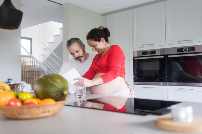 Pregnant woman and husband consulting recipe book in bright kitchen