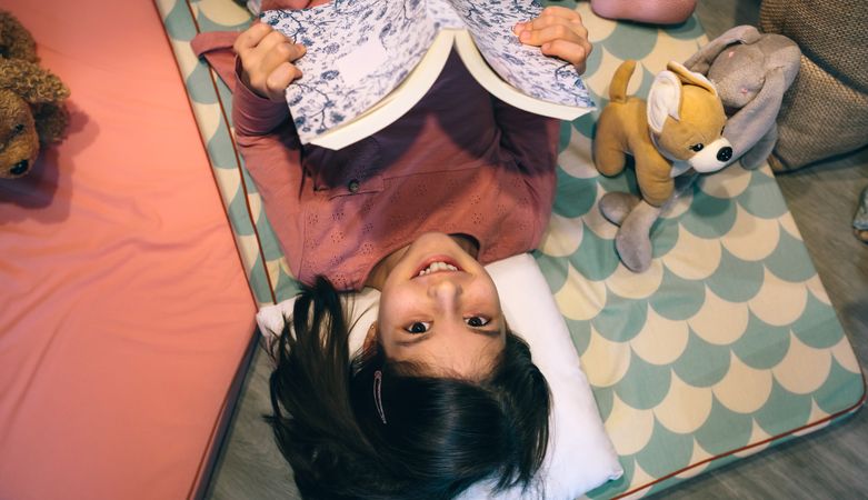 Top view of smiling little girl looking at camera while holding book and resting over sleeping mats