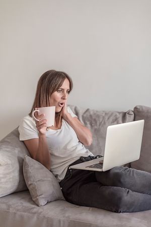 Surprised woman on sofa with a cup of coffee working on her laptop in the morning
