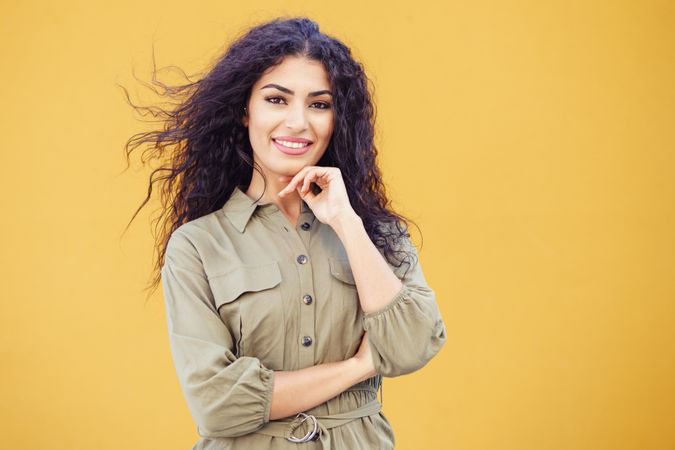 Female in army green jumpsuit smiling in front of mustard wall with hand to her chin