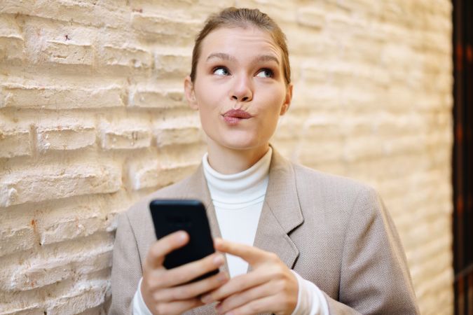 A thoughtful woman is using her smartphone while standing against a textured brick wall outside