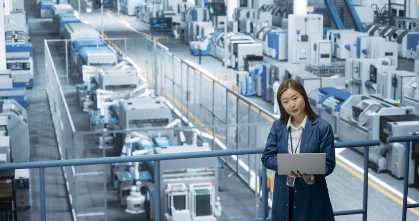 Asian Female Engineer Standing in a Factory Facility, Working on Laptop Computer, Researching and Developing Technologies to Produce Electronics at a Modern Automated Manufacture with Robotic Arms