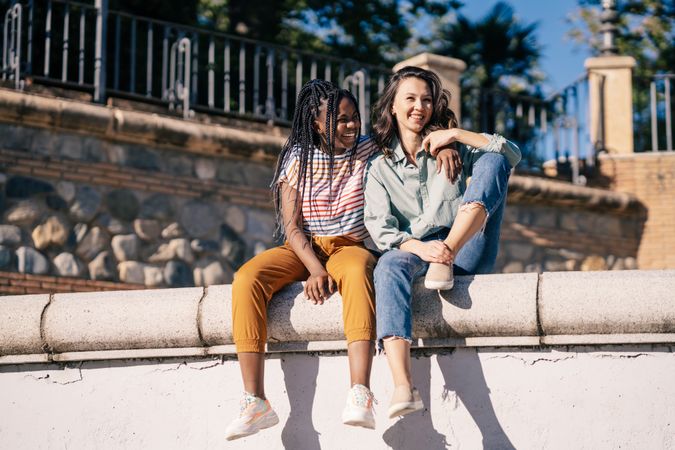 Female friends sitting on outdoor wall on sunny day