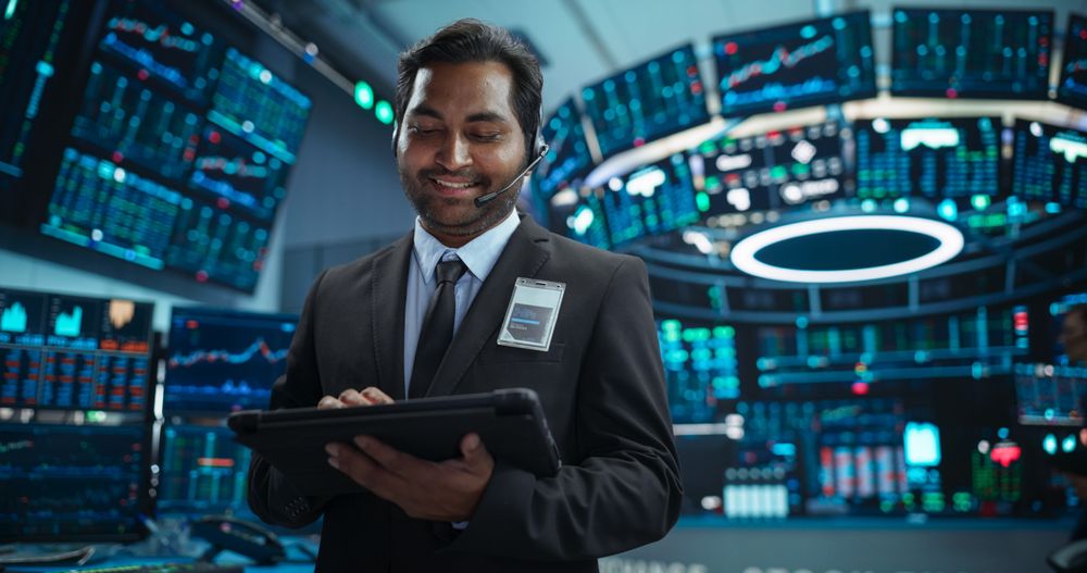 Portrait of a Young Indian Man Working in an International Stock Exchange Company: Happy Trader Using a Tablet Computer, Monitoring, Talking to a Corporate Business Partner in Headphones