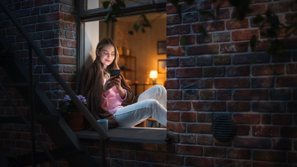 Happy White Woman Using her Smartphone While Sitting on her Windowsill During Evening.