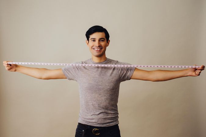 Smiling Hispanic male holding measuring tape wide between both arms in beige studio shoot