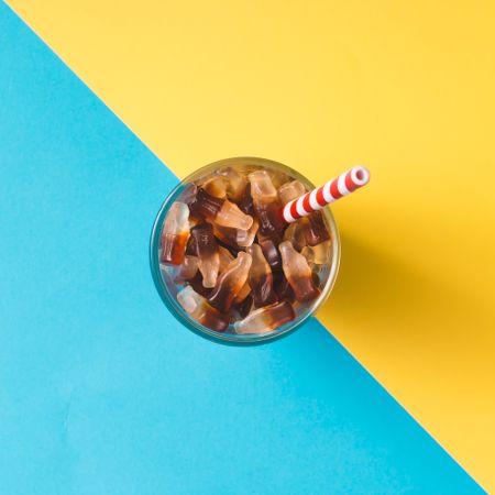 Glass full of gummy candies with straw on blue and yellow background