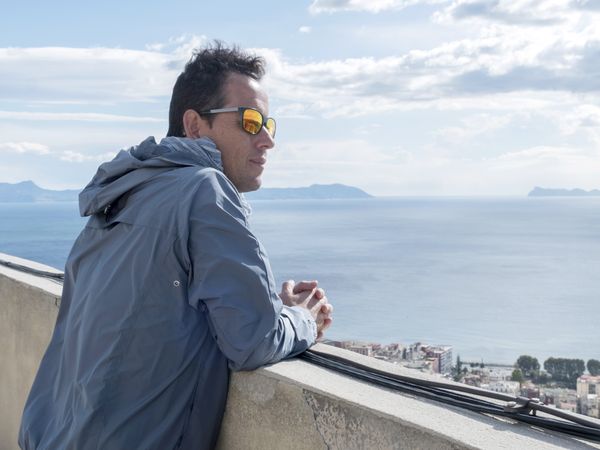 Man closeup in blue jacket leaning on railing looking away to the sea in Naples, Italy