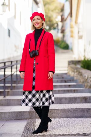 Happy adult female in elegant coat and beret with camera on neck standing on stairs