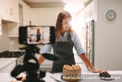 Smiling young woman standing at kitchen counter with pastries and camera bYwKX5
