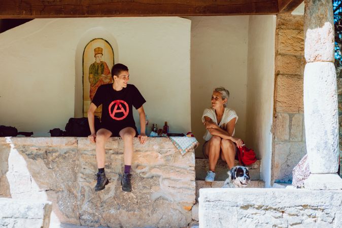 Young woman sitting on stone fence and speaking with older woman on steps of houses