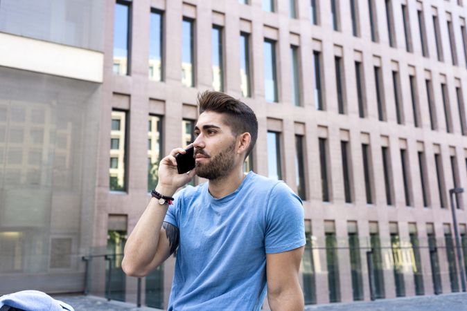 Man with beard in blue t-shirt using phone while sitting outside building