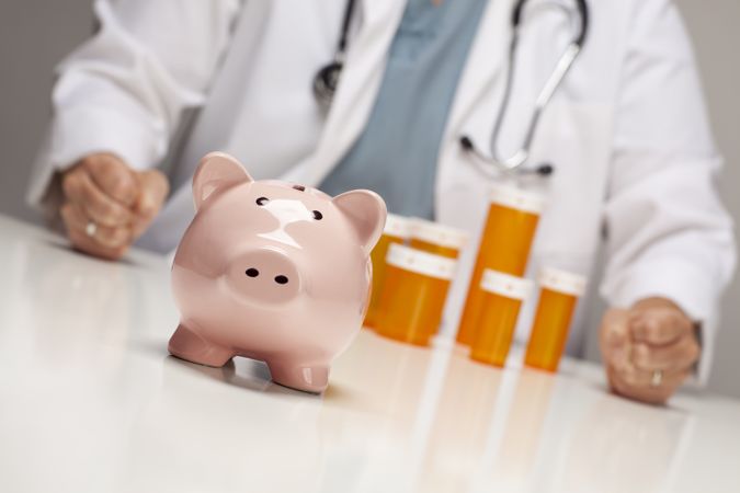 Doctor with Fists on Table Behind Bottle and Piggy Bank