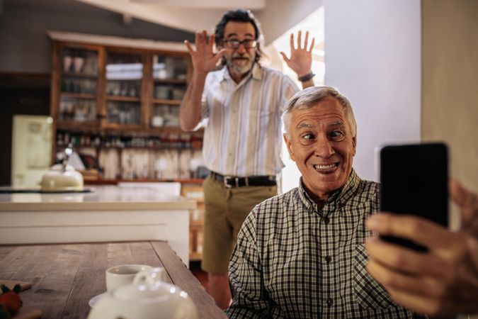 Funny man taking selfie with friend standing at back making hand gestures