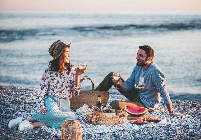 Couple smiling holding wine glasses over summer picnic by the ocean