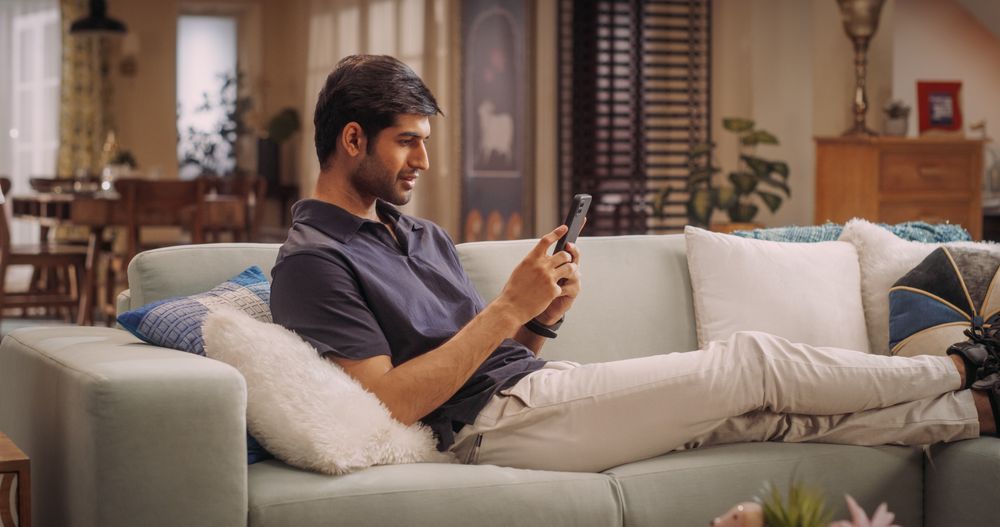 Indian Man Lying on Sofa Using Smartphone, Exploring Online Shopping, Remote Work, And Watching funny Videos