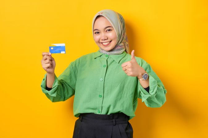 Muslim woman in headscarf and green blouse holding credit card and giving thumbs up