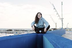 Young teenage girl sitting on a stone wall with her legs crossed while smiling at the coast