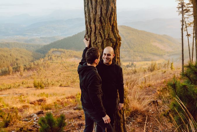 Smiling men hugging near tree in forest on hill
