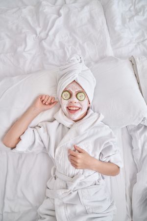 Kid in bathrobe and facial mask relaxing on bed at home