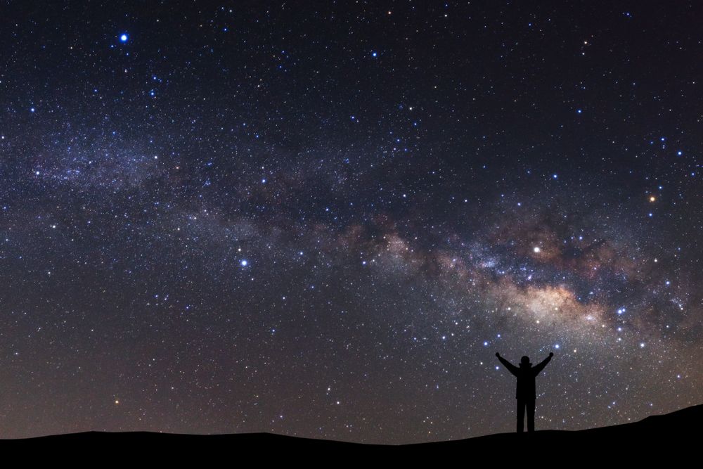 Night sky with stars and silhouette of a standing sporty man with raised up arms on high mountain.