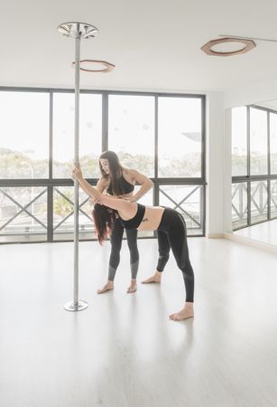 Woman helping dancer to stretch leg near pole in studio