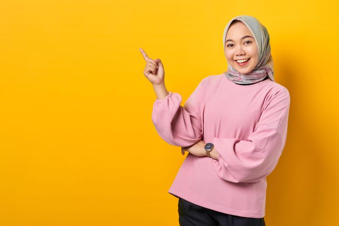 Happy Muslim woman in scarf and pink sweater pointing up at copy space studio shoot