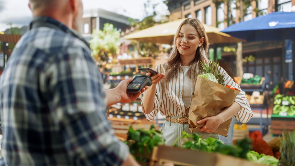 Modern Female Shopper Using Smartphone with Contactless Payment Technology to Pay for Organic Vegetables at a Farmers Market