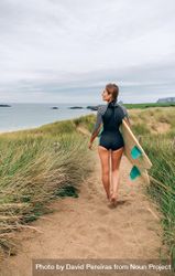 Woman walking on sandy path towards the beach holding a surfboard, vertical composition 4jKlrb