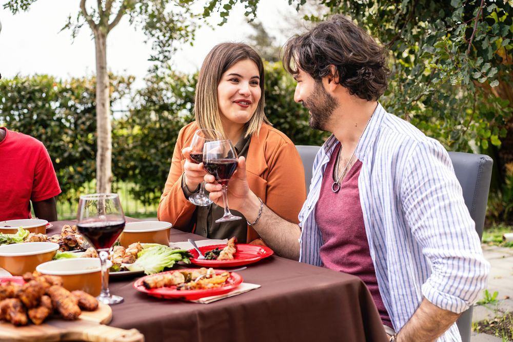 A couple engages in a friendly conversation over wine at an outdoor dining event, with a table full of various dishes in a lush garden setting