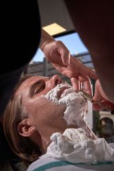 Crop barber shaving beard of adult client with razor