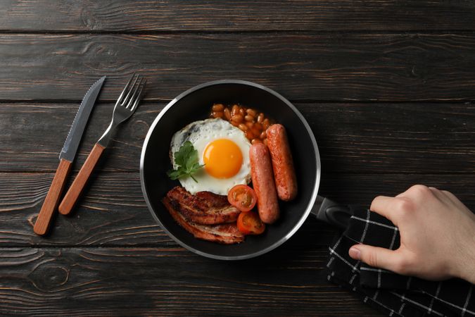 Top view of hand reaching for pan with fried breakfast of eggs, bacon and sausage