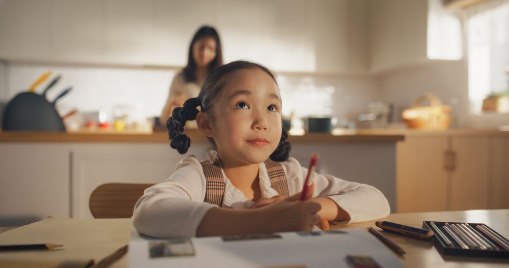 Portrait of a Little Korean Girl Sitting at a Kitchen Table at Home, Doing her Homework in the Morning While her Mother is Cooking in the Background.