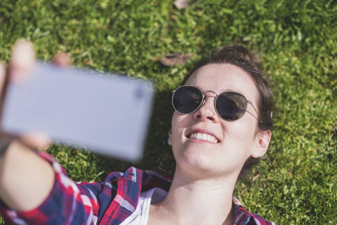 Above view of a young smiling hipster woman lying on grass in a sunny day at a park while taking a selfie with a mobile phone