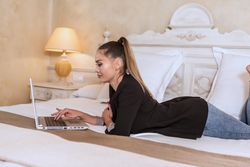 Side view of woman in dark blazer laying forward on bed using laptop