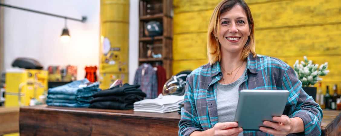 Banner of smiling blonde young female entrepreneur looking at camera in front of shop counter