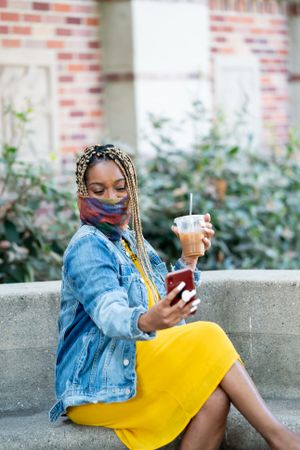 Woman sitting in front of library wearing mask taking a selfie and holding iced coffee