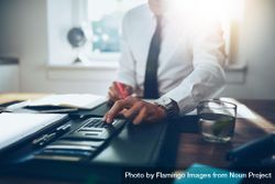 Man in shirt and tie sitting at desk with calculator in sunny office 48q6kb