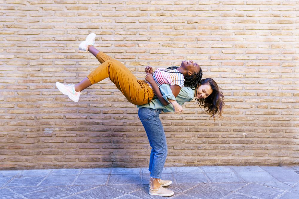 Two cheerful friends with one lifting the other up in front of brick wall