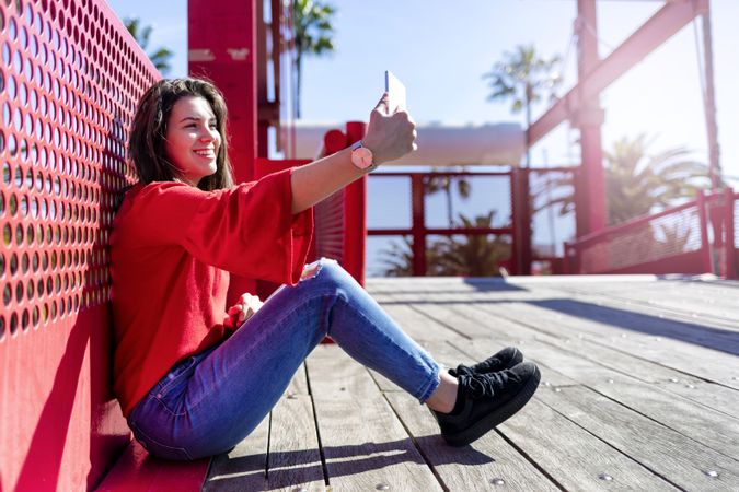 Side view of happy woman in red sitting taking selfie on mobile phone outdoors on a bright day