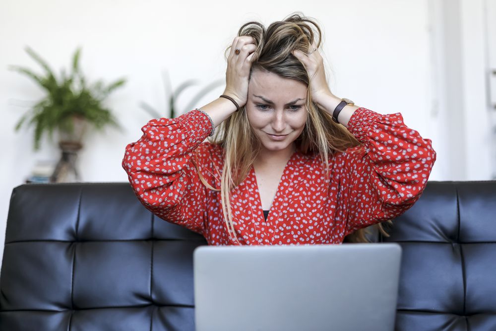 Young white woman using laptop at home sitting on the sofa scared and amazed with hands on head