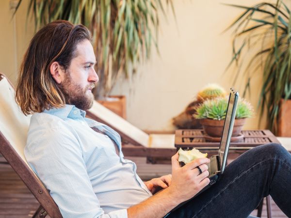Handsome male browsing laptop and enjoying apple on lounge in flat