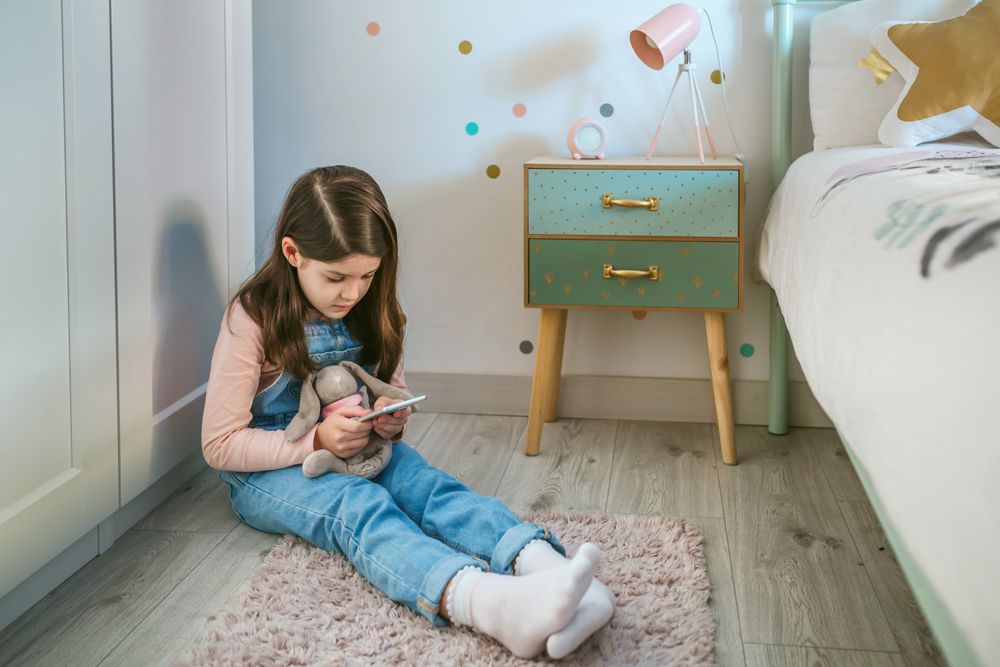 Little girl using mobile while hugging her stuffed animal sitting on the floor