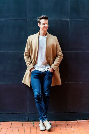 Coy man leaning on dark wall outside and wearing a camel coat