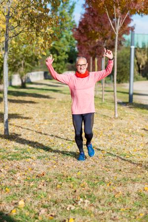 Grey haired male jogging in park with fall leaves with arms up in celebration