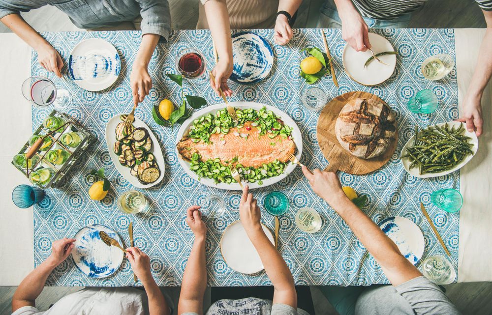 Group of people serving themselves fish, on blue patterned table with wine and side dishes and bread