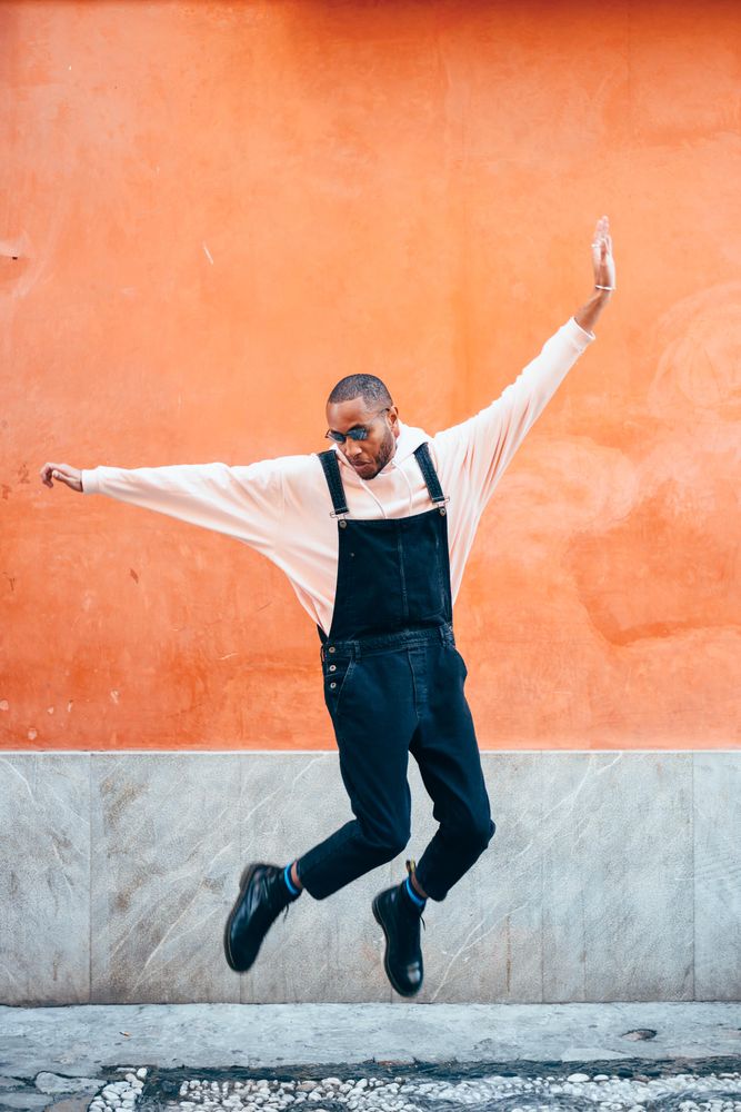 Male in overalls and hoodie jumping while looking down in front of wall