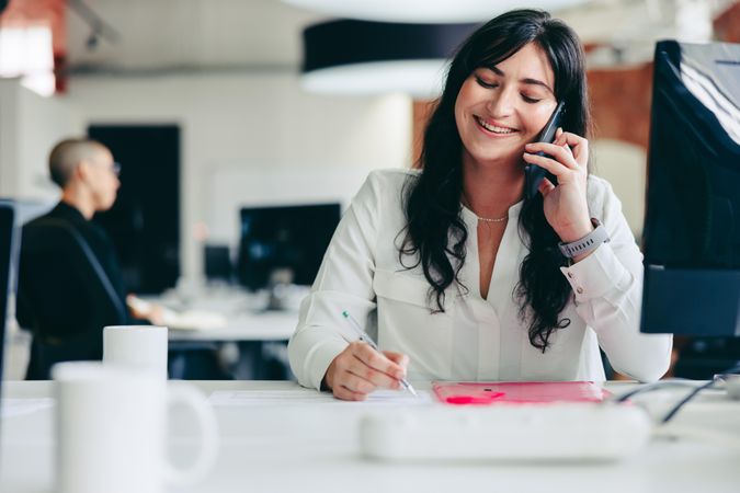 Woman at desk in modern office on phone while taking notes