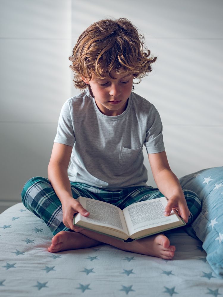 Concentrated little schoolboy reading book on bed