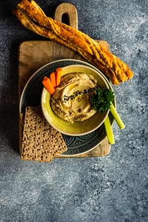 Top view of creamy hummus with bread sticks and rye crackers on grey kitchen counter